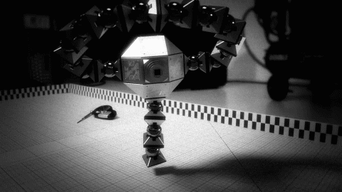 Black and white animation of a geometric robot with many cube-shaped arms, standing on a tiled floor.